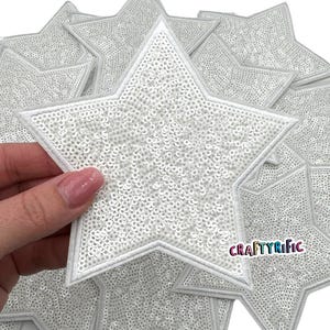 May include: A pack of white star-shaped iron-on patches with a sparkly sequin finish. The patches are approximately 3 inches in diameter. The text "CRAFTYrific" is visible in the bottom right corner of the image.