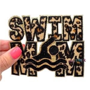 May include: A rectangular patch with the word "SWIM" in large, leopard print letters. A black swimmer graphic is in the center, with gold glitter accents. The patch is held by a hand with pink nail polish.