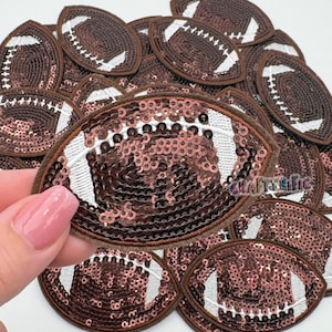 May include: A close-up of a pile of brown and white football-shaped patches with sequins. The patches are made of brown fabric with white stitching and have a white sequin design.