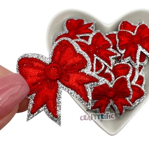 May include: A close-up of a red glitter bow with a silver glitter trim. The bow is on a white heart-shaped surface. There are many other red glitter bows with silver glitter trim in the background.