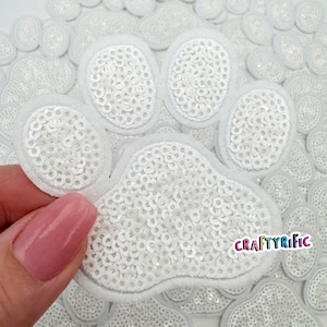 May include: White paw print shaped sequins iron-on patch. The patch is made of white sequins sewn onto a white fabric base. The patch is surrounded by other paw print shaped sequins.