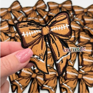 May include: A close-up of a brown and white football-themed bow. The bow is made of fabric and has a stitched design that resembles a football. The bow is surrounded by other similar bows.