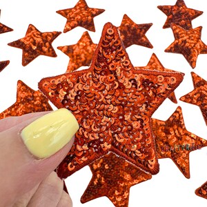 May include: A close-up of a hand holding a large orange star-shaped sequin patch. The patch is made of small, shiny sequins and has a felt backing. There are several other smaller orange star-shaped sequin patches in the background.
