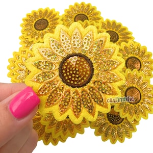May include: Embroidered yellow sunflower patches with gold sequins and brown centers. The patches are in a pile, with one being held by a hand with pink nail polish. The patches are from CraftyRific.