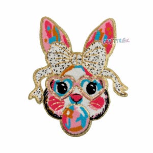 May include: Embroidered patch featuring a bunny face with heart-shaped sunglasses and a bow. The patch has a white, pink, blue, and gold color scheme. The text "CraftyRific" is visible on the right ear.