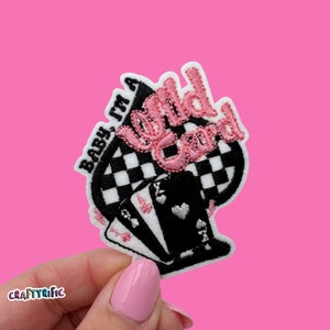 May include: Embroidered patch with the text "Baby, I'm a Wild Card" in black and pink. The patch features a black and white checkered background and three playing cards. The patch is held against a pink background.