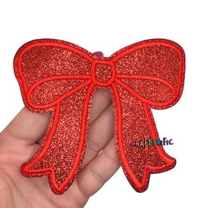 May include: A red glitter bow applique with a stitched outline. The bow has a classic bow shape with two loops and two streamers. The text "CRAFTYRIFIC" is visible on the right side of the bow.