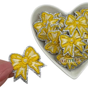 May include: A pile of yellow and silver glitter bow appliques in a heart-shaped dish. The bows are about 1 inch in size and have a silver glitter outline.