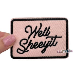 May include: A rectangular pink patch with a black border. The patch features the embroidered phrase "Well Sheeeyit" in black cursive lettering. The lower right corner has the text "CRAFTYRIFIC".