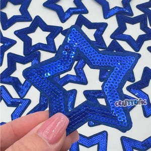 May include: A close-up of a hand holding a blue sequined star-shaped patch. The patch is surrounded by other blue sequined star-shaped patches.