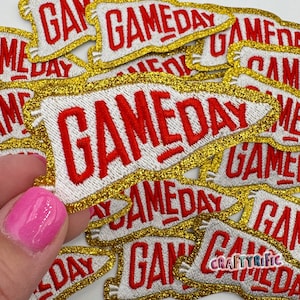 May include: White and gold embroidered "GAMEDAY" patches with red lettering. The patches have a gold glitter border and are in a pennant shape. The patches are for sports fans.