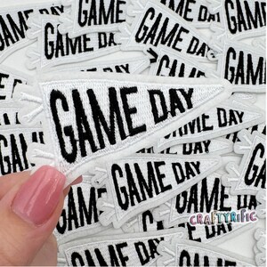 May include: A close-up of a pile of white felt pennant patches with black embroidered text that reads "GAME DAY".