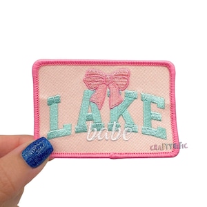 May include: A rectangular patch with a pink border and a light peach background. The patch features the words "LAKE babe" in teal and white embroidery, with a pink bow above the word "LAKE". The text "CRAFTYETIC" is in the bottom right corner.