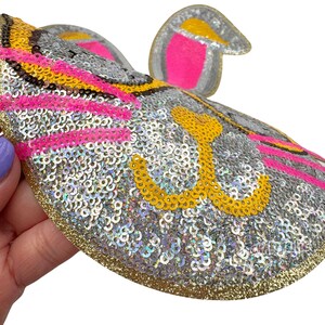 K&ouml;nnte beinhalten: Ein glitzernder, silberner, rosa und gelber Pailletten-Aufn&auml;her in Form eines Tierkopfes aus einem Cartoon. Der Aufn&auml;her hat einen goldenen Rand.