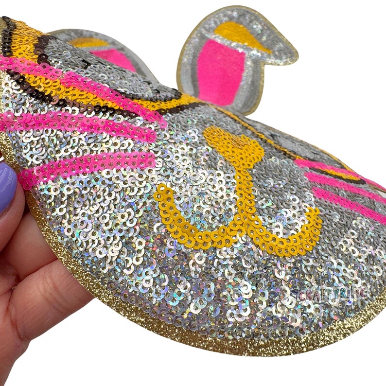 K&ouml;nnte beinhalten: Ein glitzernder, silberner, rosa und gelber Pailletten-Aufn&auml;her in Form eines Tierkopfes aus einem Cartoon. Der Aufn&auml;her hat einen goldenen Rand.