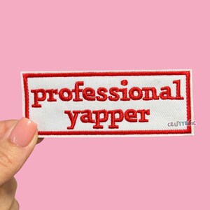 May include: A rectangular white patch with a red border and the words "professional yapper" embroidered in red. The patch is held against a pink background.
