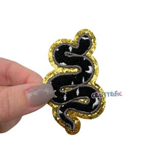 May include: A black snake with white markings is embroidered on a gold glitter iron-on patch. The patch is shaped like the snake and has a gold glitter outline.