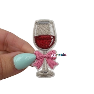 May include: Embroidered patch featuring a wine glass filled with red liquid, adorned with a pink bow. The patch has a gray outline and the text "CRAFTYrific" is visible on the bow.