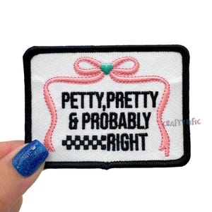May include: A rectangular patch with a white background and black border. The patch features the text "PETTY, PRETTY & PROBABLY RIGHT" in black, with a pink ribbon design and a teal heart. The patch is held by a hand with a blue glitter nail polish.