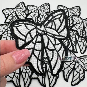May include: A set of black and white embroidered iron-on patches in the shape of a bow with a spiderweb design.