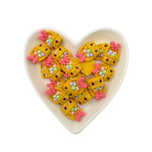 May include: A collection of yellow, smiling, basket-shaped charms with pastel-colored candies inside. The charms have pink bows and are arranged in a heart-shaped dish.