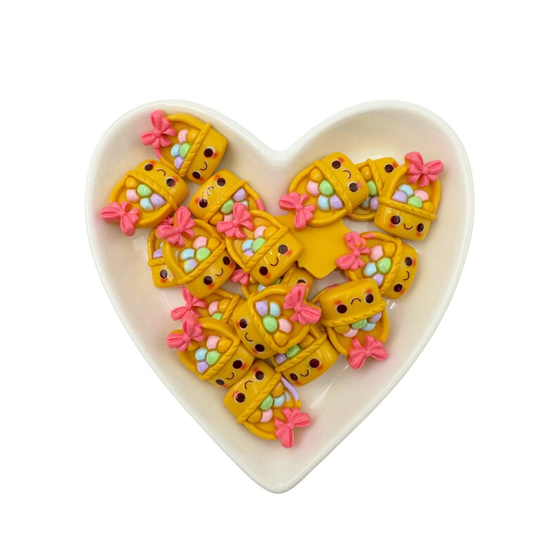 May include: A collection of yellow, smiling, basket-shaped charms with pastel-colored candies inside. The charms have pink bows and are arranged in a heart-shaped dish.