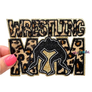 May include: A rectangular patch with the words "WRESTLING MOM" in a gold glitter outline. The letters "WRESTLING" have a leopard print pattern. The word "MOM" has the same leopard print pattern. A black glitter silhouette of two wrestlers is in the center.