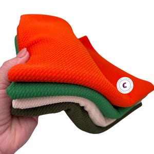 K&ouml;nnte beinhalten: Vier St&uuml;cke Waffelstrickstoff in den Farben Orange, Gr&uuml;n, Beige und Olivgr&uuml;n. Der Stoff ist gefaltet und wird in einer Hand gehalten.