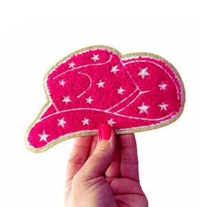 May include: A hot pink cowboy hat patch with white star accents and a gold glitter border. The patch is a fun accessory for jackets, bags, or other fabric items. The patch is approximately 4 inches wide.