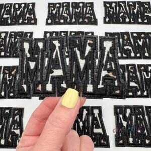 May include: A close-up of a hand holding a black and white "MAMA" patch with a leopard print design. The patch is surrounded by other similar patches.