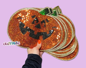 May include: A stack of orange sequined pumpkin-shaped placemats with gold trim and green leaf accents. Each pumpkin has a smiling jack-o'-lantern face with black sequin details. The placemats are held up against a light purple background.