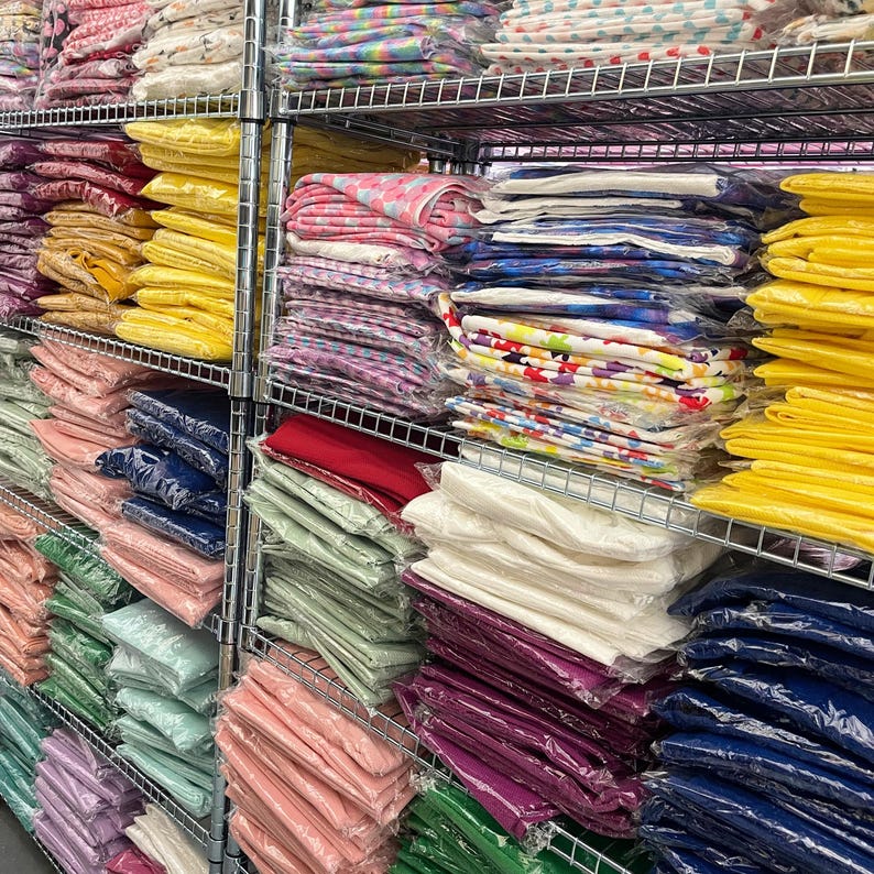 K&ouml;nnte beinhalten: Ein Metallregal gef&uuml;llt mit Stapeln bunter Stoffe. Der Stoff ist gefaltet und in Plastik verpackt. Die Regale sind gef&uuml;llt mit verschiedenen Farben und Mustern, darunter Rot, Wei&szlig;, Blau, Gr&uuml;n, Gelb und Rosa.