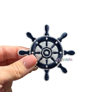 May include: Embroidered patch of a ship's wheel in navy blue and white. The nautical-themed patch features a detailed design with a circular shape and six spokes. The patch is held by a person's hand.