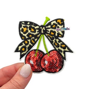 May include: Embroidered patch featuring two red sequined cherries with green stems, topped with a leopard print bow. The patch has a white border and the text "CRAFTYRIFIC" is visible.