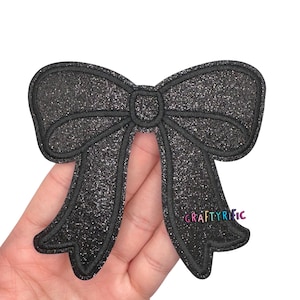 May include: A black glitter bow applique with a stitched outline. The bow has a classic bow shape with two loops and two streamers. The text "CRAFTYRIFIC" is visible on the right side of the bow.