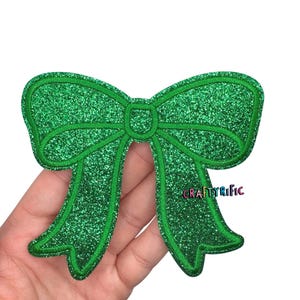 May include: A green glitter bow with a stitched outline. The bow is held in a hand. The text "CRAFTYrific" is visible on the bow. This decorative bow is suitable for crafts and embellishments.