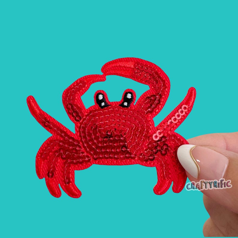 May include: A bright red crab-shaped patch covered in shimmering sequins. The crab features two raised claws, multiple legs, and black and white eyes. The text CRAFTYRIFIC is visible in small white letters on the right claw and near the edge.