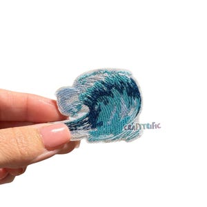 May include: Embroidered patch featuring a stylized ocean wave in shades of blue and white. The patch has a detailed, textured appearance, with the text "CRAFTYRIFIC" visible on the lower right. Ideal for adding to clothing or accessories.