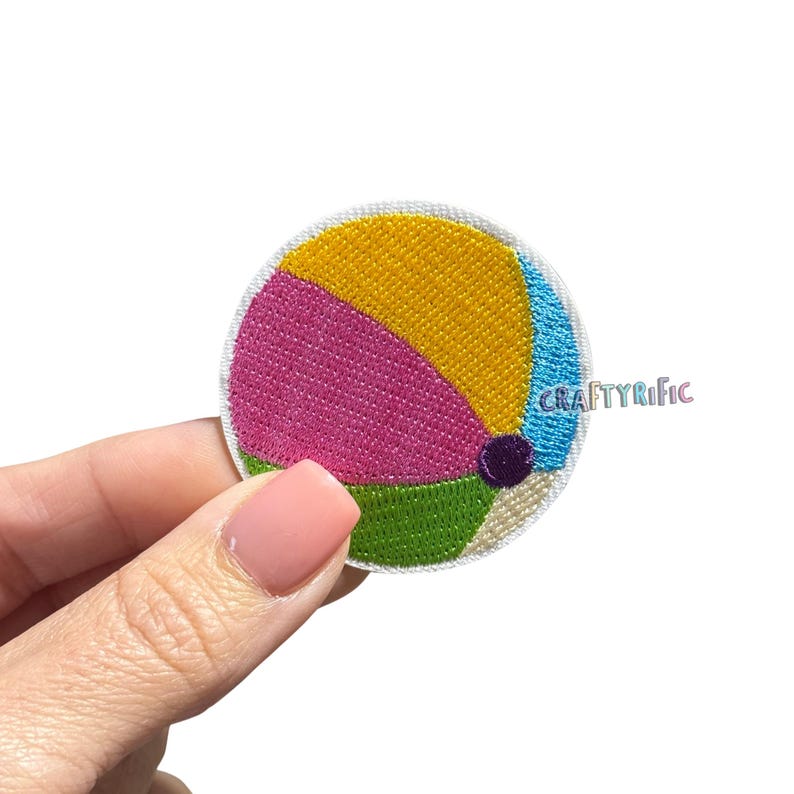 May include: Embroidered patch in the shape of a beach ball. The patch features segments of yellow, pink, green, and blue, with a small purple dot. The text "CRAFTYRIFIC" is visible on the right side.