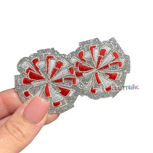 May include: Two glittery silver cheerleading pom-pom patches with red and white accents. The patches are embroidered with a starburst design. The text "CRAFTYRIFIC" is visible on the right side.