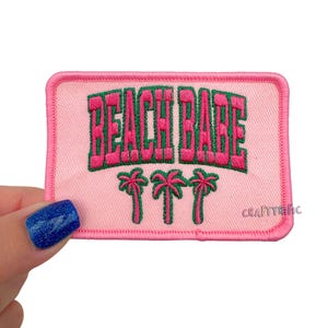 May include: Pink embroidered patch with the words "BEACH BABE" in pink and green, with three palm trees below. The patch has a pink border and is rectangular in shape.