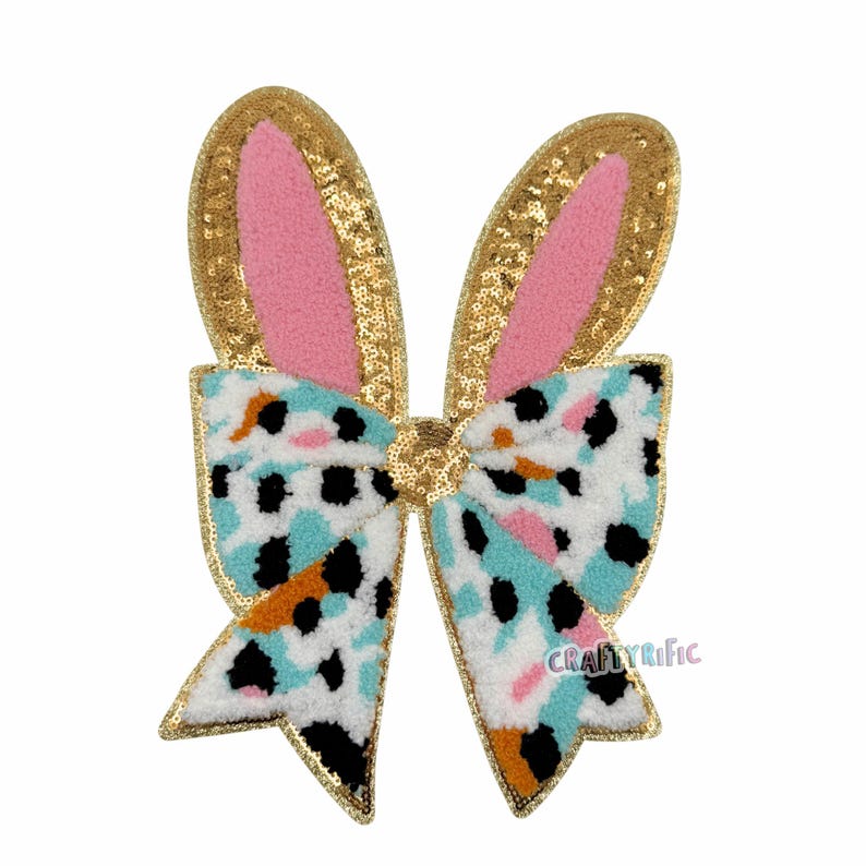May include: A decorative Easter bow featuring bunny ears. The ears are gold sequined with pink inner ears. The bow has a white, blue, black, and orange pattern, outlined in gold sequins. Ideal for Easter celebrations.
