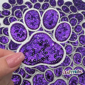 May include: Purple sequin paw print applique with a white felt backing. The paw print is surrounded by other smaller purple sequin paw prints.