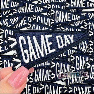 May include: A close-up of a hand holding a blue felt pennant with white embroidered text that reads "GAME DAY". The pennant is surrounded by other similar pennants.
