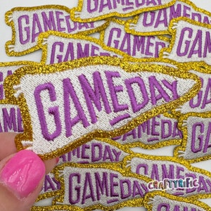 May include: A close-up of a pile of iron-on patches with a gold glitter border and purple embroidery that reads "GAMEDAY".