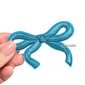 May include: A teal embroidered bow patch with a detailed stitch design. The patch has a classic bow shape with loops and a center knot. The text "CRAFTYrific" is visible on the lower right side of the bow.