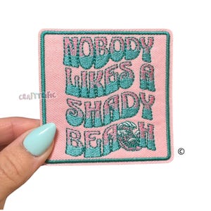 May include: A pink embroidered patch with the text "NOBODY LIKES A SHADY BEACH" in teal lettering. The patch has a teal border and is held by a hand with a light blue nail polish.