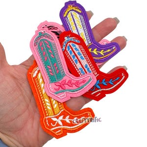 May include: A collection of colorful embroidered cowboy boot patches in orange, pink, red, and purple. Each boot features intricate floral and geometric designs. The orange boot has the text "CraftyRific".