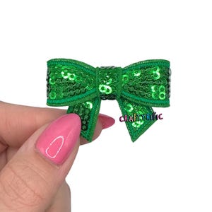 May include: A small, green sequined bow with a stitched edge. The bow is held in a hand with pink nail polish. The text "CRAFTYrific" is visible on the bow.