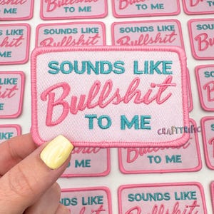 May include: A rectangular pink patch with the phrase "SOUNDS LIKE Bullshit TO ME" embroidered in teal and pink. The patch has a white background and a pink border. The word "CraftyRific" is printed on the bottom right.